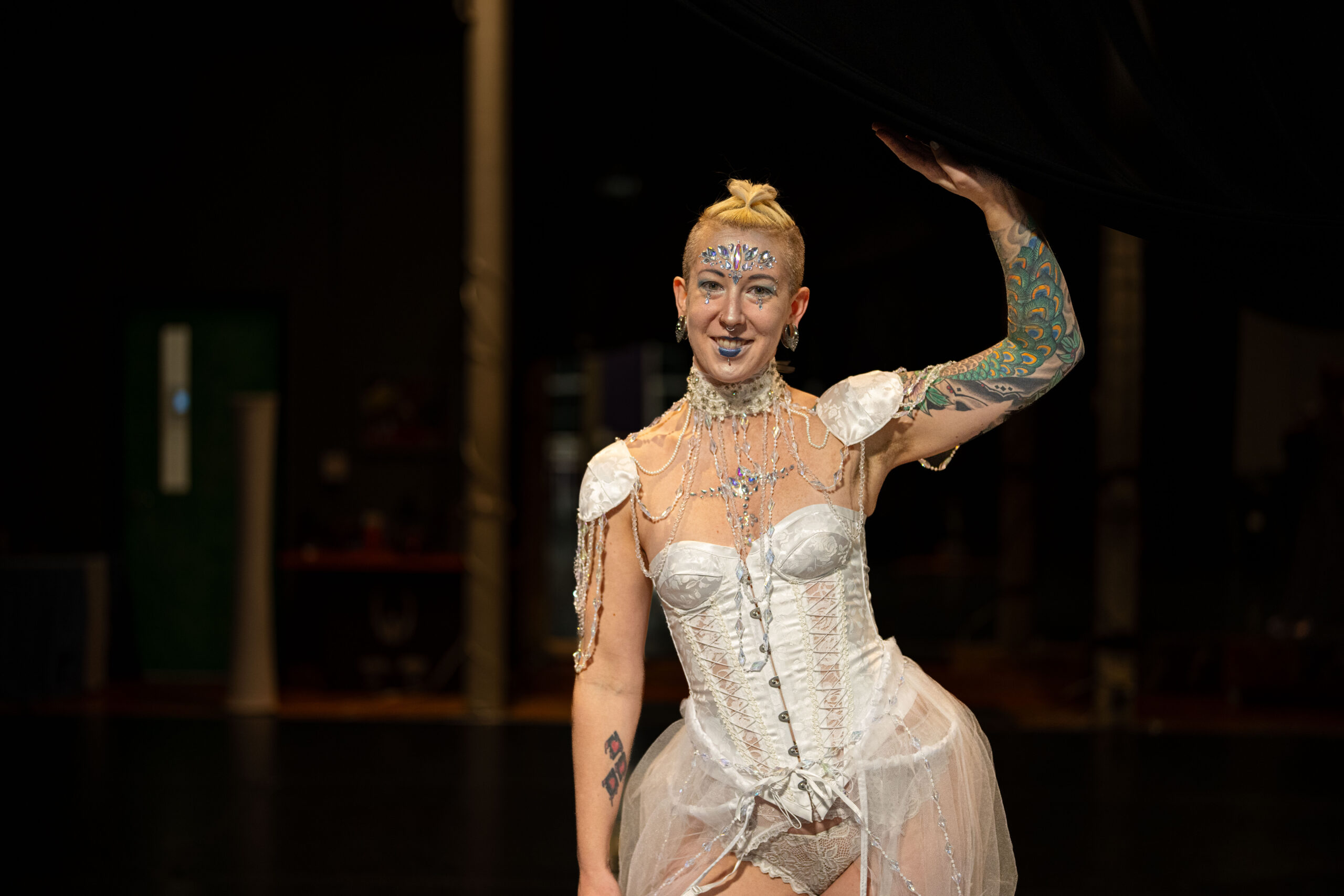 A woman with tattoos and blond hair in a white burlesque costume standing backstage.