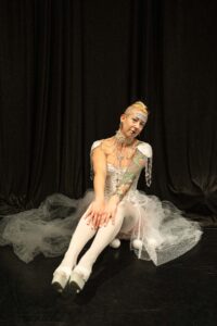 A white woman with tattoos and blonde hair sitting in a white burlesque costume.