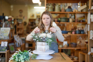 a white, blond woman arranging white flowers in a store.