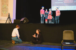 Two individuals sitting on the floor front of a stage signing the lyrics to the music being played on stage.