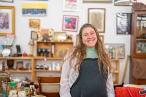 A white woman in an apron standing in front of art mounted on the wall of a store.