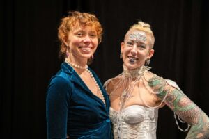 Two white women smiling. One with red hair and a blue velvet blazer. One with blonde hair, a white corset, jewels on her forehead, and tattoos on her arm.