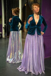 A white woman with red hair wearing a purple dress and blue velvet blazer. A mirror reflects her back.