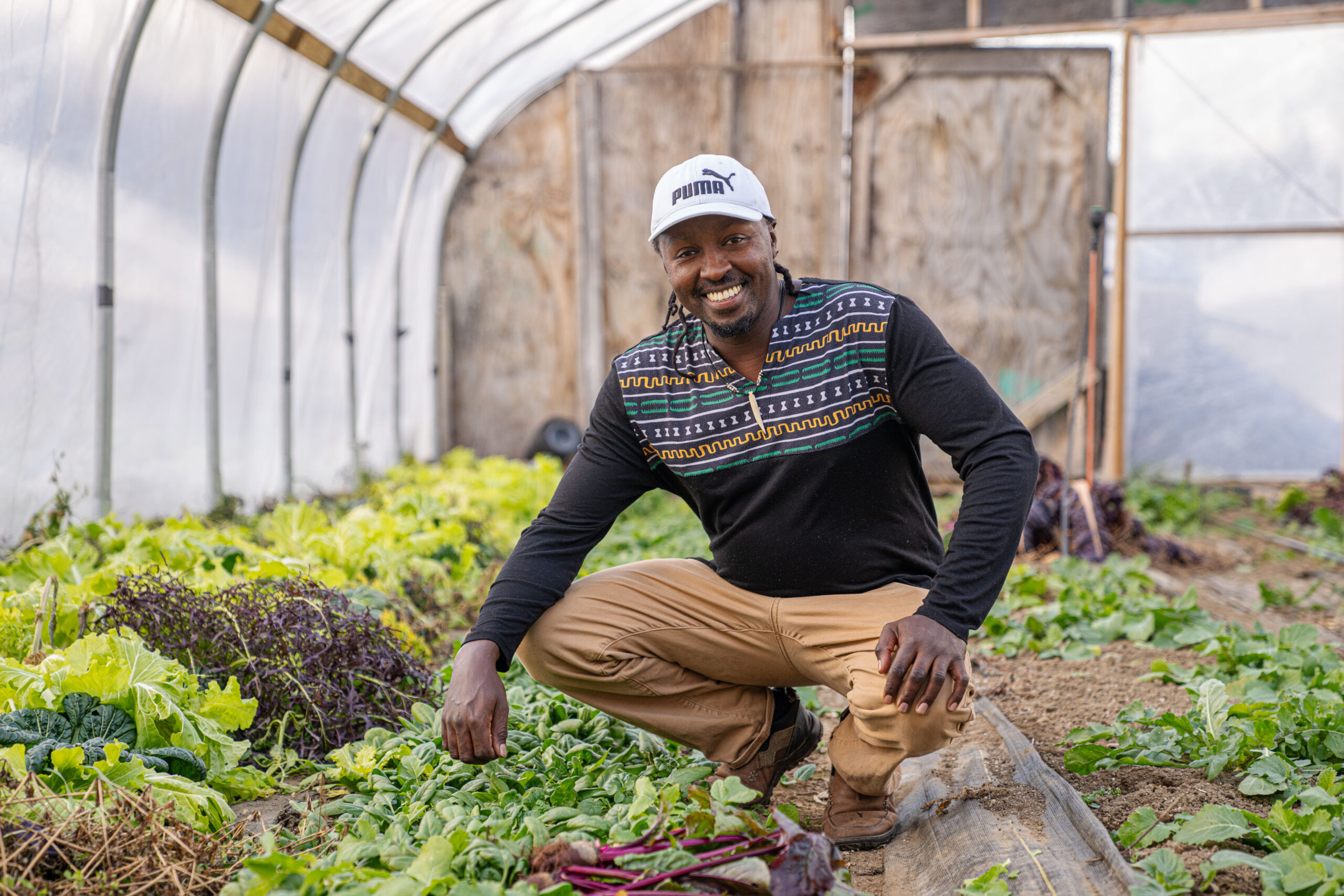 A Black man in a greenhouse crouching near his vegetables.