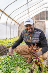 A Black man crouching in a greenhouse, holding a bunch of beets.